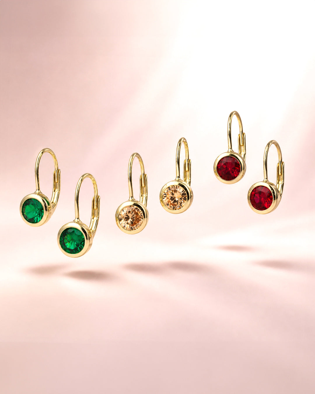 Golded leverback earrings with round bezel-set stones in green, copper, and red, arranged on a soft pink gradient background.