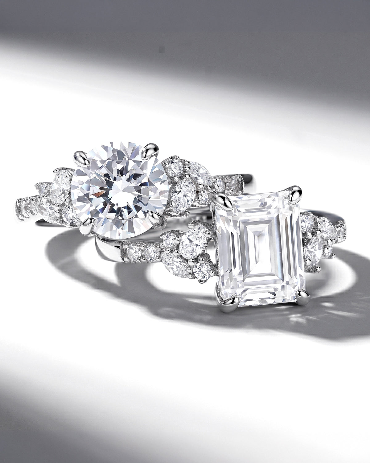 A close-up studio shot of a silver-toned rings with multiple clear stones, including a large round brilliant-cut center stone and an emerald-cut stone, photographed on a white background with dramatic shadows.